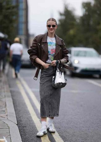 Die Frau trägt eine braune Lederjacke, ein grafisches T-Shirt, einen Jeansrock und Adidas-Turnschuhe