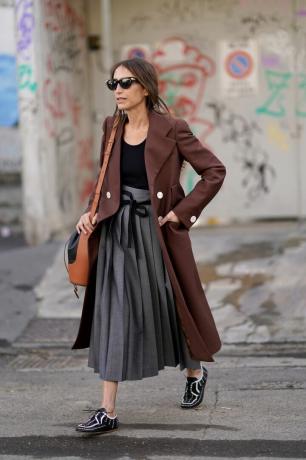 Chloé Harrouche porte du marron et du noir avec une veste longue et un haut noir avec une jupe fluide.