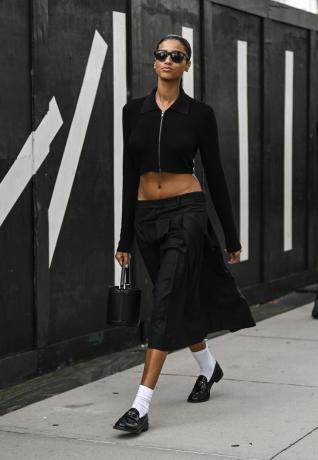 Žena jdoucí po chodníku v černých slunečních brýlích, v černém tričku s dlouhým rukávem a v černé sukni