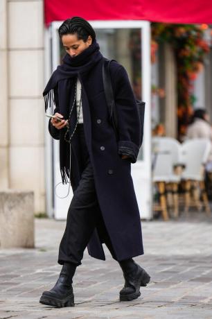 Жінка в чорних черевиках Челсі з чорними брюками та чорним пальто з великим чорним шарфом. 