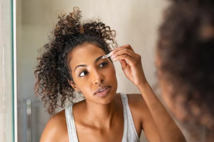 Afroamerikansk kvinna som plockar ögonbryn med pincett medan hon står framför spegeln.