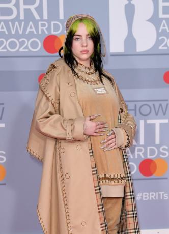 Billie Eilish su smėlio spalvos „Burberry“ paltu, dideliu viršutiniu dydžiu ir kelnėmis su auksinės grandinėlės detalėmis