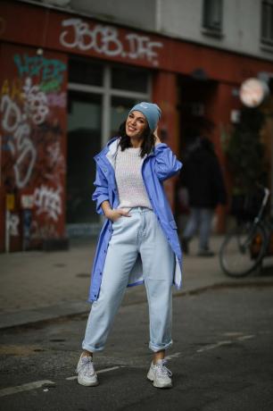 Anna Wolfers trägt einen weißen Strickpullover, Jeans in heller Waschung, eine lila Jacke, eine blaue Mütze und weiße Turnschuhe