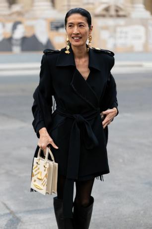Žena v černém kabátě se zlatými visacími náušnicemi a zlato-béžovou hranatou taškou