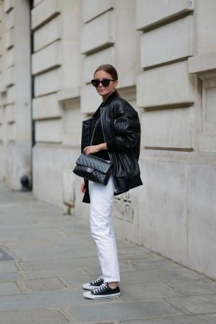 Женщина носит огромную кожаную куртку с белыми брюками и конверсами.