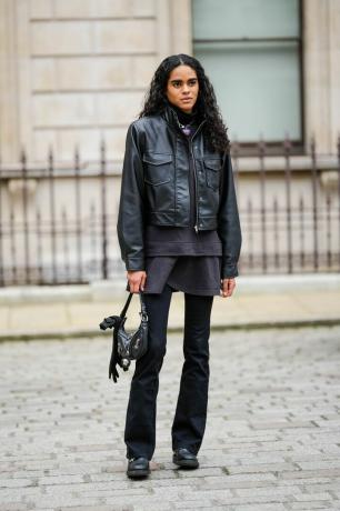 Modelka v černém dlouhém tričku, černém svetru, černé lesklé kožené bundě, černých džínových kalhotách, v ruce černou lesklou koženou tašku.