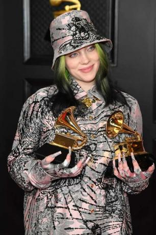 Billie Eilish con un sombrero de pescador estampado en rosa claro, negro y plateado, atuendo y uñas sosteniendo dos premios Grammy